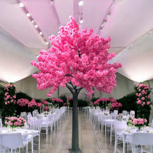 Load image into Gallery viewer, A stunning artificial pink cherry blossom tree is prominently displayed in the center of an elegant event venue. The room features white chairs and tables adorned with pastel floral arrangements, set against a backdrop of lush greenery and a high, translucent ceiling that allows soft natural light to fill the space.