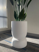 Charger l'image dans la galerie, Close-up of a white stackable planter with a smooth matte finish, holding a tall artificial plant with large green leaves, placed indoors near a window with natural light. Does this planter have drainage holes?