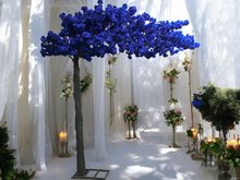 Load image into Gallery viewer, A vibrant blue artificial tree is set against an ethereal backdrop of flowing white drapery in an elegant venue. The space is adorned with floral arrangements in soft pastel tones, complemented by golden stands and glowing candles, creating a romantic and serene ambiance.