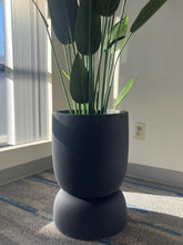 Charger l'image dans la galerie, Close-up of a black stackable planter with a smooth matte finish, holding a tall artificial plant with large green leaves, placed indoors near a window with natural light. Does this planter have drainage holes?