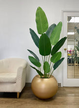 Load image into Gallery viewer, Artificial bird of paradise plant with vibrant green leaves in a large gold spherical planter placed beside a cream-colored armchair in a bright, modern indoor space