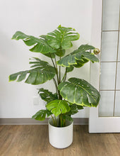 Load image into Gallery viewer, Artificial monstera plant in a white ceramic pot with a wooden stand, positioned next to a cream-colored armchair. The plant features large, realistic green leaves with natural splits, adding a tropical vibe to the indoor space. The setup is placed on a carpeted floor with subtle blue accents