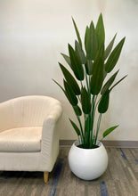 Load image into Gallery viewer, An artificial bird of paradise plant with tall green leaves in a round white pot. The plant is placed next to a white, cushioned chair, adding a natural touch to the room's modern decor. The clean and minimalist setting emphasizes the plant's vibrant and realistic appearance