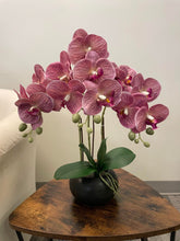 Charger l'image dans la galerie, A beautiful arrangement of pink artificial orchids in a black planter, placed on a wooden side table. Frequently asked question: What are the best care tips for maintaining artificial orchids?
