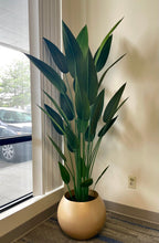 Load image into Gallery viewer, A tall artificial bird of paradise plant in a gold round planter positioned by a large window. The plant features realistic, elongated green leaves, adding a touch of natural elegance to the modern indoor space. The room's neutral tones highlight the vibrant green of the plant, making it an eye-catching decor element