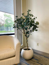 Load image into Gallery viewer, Artificial plant with broad, green leaves displayed in a modern, round white pot, placed next to a beige armchair, adding a natural touch to the indoor setting.
