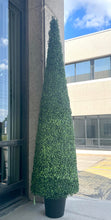 Charger l'image dans la galerie, A tall, artificial topiary shaped like a cone stands in a black pot outside a building with large glass windows. The topiary is covered in small, tightly packed green leaves. The reflection of trees and a cloudy sky is visible in the glass behind it.