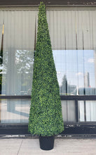 Charger l'image dans la galerie, A tall, artificial topiary in the shape of a cone stands in a black pot. The topiary is lush with dense, small green leaves and is placed outside against a textured concrete wall. In the background, there is a view of a building with large windows