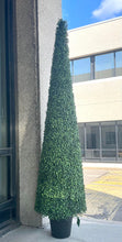 Charger l'image dans la galerie, A tall, artificial topiary shaped like a cone stands in a black pot outside a building with large glass windows. The topiary is covered in small, tightly packed green leaves. The reflection of trees and a cloudy sky is visible in the glass behind it