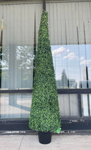 Charger l'image dans la galerie, A tall, artificial topiary in the shape of a cone stands in a black pot on a gray carpet. The topiary is positioned next to a white upholstered chair, providing a contrast between the soft texture of the chair and the dense, leafy surface of the topiary