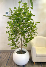 Load image into Gallery viewer, Artificial tree in a round white planter placed next to a cream-colored chair, enhancing the indoor space with natural-looking greenery.