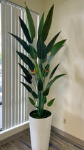 Load image into Gallery viewer, Tall artificial bird of paradise plant with dark green leaves and bright orange flowers, placed in a large white textured planter. The plant is positioned near a window with vertical blinds, enhancing the natural light in the room and adding a tropical touch to the indoor space.