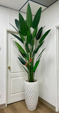 Load image into Gallery viewer, Tall artificial bird of paradise plant with large green leaves and vibrant orange flowers, set in a textured white ceramic planter. The plant is positioned indoors against a white wall and door, adding a touch of greenery to the space.