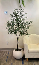 Load image into Gallery viewer, Elegant living room setup featuring a tall artificial olive tree in a white pot positioned next to a modern beige sofa with gold trim. The room has a warm, brown-toned wall adorned with a colorful landscape painting, creating a cozy and sophisticated atmosphere.