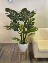 Charger l'image dans la galerie, Artificial monstera plant in a white ceramic pot with a wooden stand, positioned next to a cream-colored armchair. The plant features large, realistic green leaves with natural splits, adding a tropical vibe to the indoor space. The setup is placed on a carpeted floor with subtle blue accents."