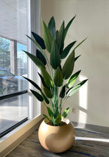 Load image into Gallery viewer, Tall artificial bird of paradise plant with lush green leaves placed in a round, golden planter by a large window, with natural light streaming in, enhancing the vibrant and realistic appearance of the plant.