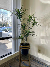 Charger l'image dans la galerie, Artificial yucca plant in a black planter with gold metal stand, displayed by a large window for modern office or home decor