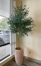 Load image into Gallery viewer, Artificial olive tree placed in a textured, light brown pot with a leaf pattern. The tree is positioned near a window with vertical blinds, filling the corner of a room with a neutral wall color and carpeted floor.