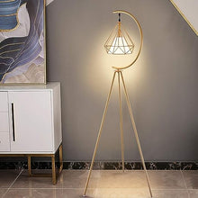 Charger l'image dans la galerie, A modern gold floor lamp with a geometric lampshade and crescent-shaped frame, placed next to a white and gold console. The lamp illuminates a soft glow against a dark grey wall, complementing the elegant, minimalist decor of the space.