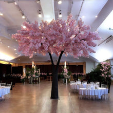 Load image into Gallery viewer, Artificial tree adorned with soft pink blossoms, beautifully displayed as a centerpiece in an elegantly decorated event hall with round dining tables and floral arrangements. Can these rental trees enhance the ambiance of formal events like weddings and banquets?