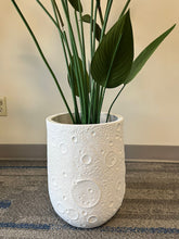 Charger l'image dans la galerie, White planter with a crater-like textured design, holding an artificial bird of paradise plant with vibrant green leaves and orange flowers, placed indoors on a carpeted floor. Is this planter made from eco-friendly materials?