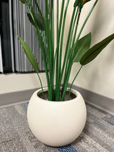 Charger l'image dans la galerie, Close-up of an ivory round planter with a smooth matte finish, holding an artificial palm plant with vibrant green leaves, placed indoors near a window. Does this planter have a built-in drainage system?