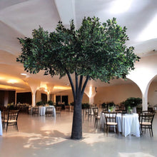 Load image into Gallery viewer, Large artificial tree with a textured trunk and lush green foliage, displayed indoors in a spacious event hall with round tables and elegant decor. Is the base of the tree adjustable for stability on different surfaces?
