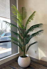 Charger l'image dans la galerie, A stylish artificial palm tree placed in a white, round planter, adding a touch of greenery and elegance to this office or living space near a window. The natural light accentuates the realistic appearance of the lush, green fronds, creating a serene and inviting atmosphere.
