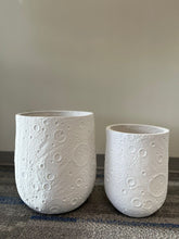Charger l'image dans la galerie, Set of two white planters with a crater-like textured design, displayed empty indoors on a carpeted floor. What are the available sizes for these planters?
