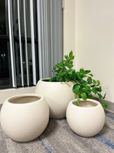 Charger l'image dans la galerie, Set of three round ivory planters with a smooth matte finish, displayed indoors near a window. One planter features a trailing artificial green plant for decoration. Are these planters available in other colors?