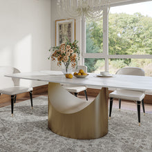 Charger l'image dans la galerie, Elegant oval marble dining table with unique golden crescent-shaped base, set with cream chairs and benches. Fresh floral arrangement and lemons adorn the table, bathed in natural light from large windows overlooking a lush garden