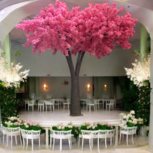 Load image into Gallery viewer, Artificial tree with vivid pink blossoms, elegantly positioned on a raised stage in a wedding venue surrounded by floral arrangements and white seating. Can these rental trees be used to create a dramatic focal point for ceremonies?