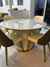 Charger l'image dans la galerie, Modern Round Dining Table with a Glossy White Top and Pleated Base, Paired with Beige and Grey Upholstered Chairs Featuring Gold Legs in a Contemporary Home Setting with Large Windows
