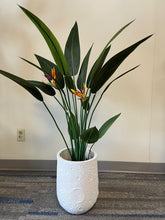 Charger l'image dans la galerie, White planter with a crater-like textured design, holding an artificial bird of paradise plant with vibrant green leaves and orange flowers, placed indoors on a carpeted floor. Is this planter made from eco-friendly materials?