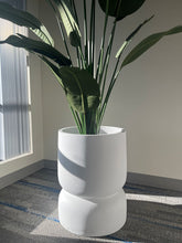 Charger l'image dans la galerie, Close-up of a white stackable planter with a smooth matte finish, holding a tall artificial plant with large green leaves, placed indoors near a window with natural light. Does this planter have drainage holes?