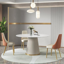 Charger l'image dans la galerie, Contemporary dining room featuring a stylish round table with a fluted base and white marble top, surrounded by four elegant chairs with beige and orange upholstery and gold legs. The setup includes modern pendant lights, gold accents, and a touch of greenery for a sophisticated look.