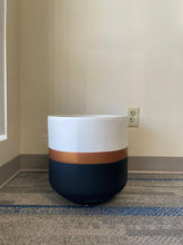 Charger l'image dans la galerie, A modern, rounded planter with a matte white top, a copper stripe in the middle, and a dark navy base sits on a gray and blue carpet near a beige wall with an electrical outlet in the background.