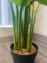 Load image into Gallery viewer, Close-up of the base of an artificial monstera plant showing the green stems emerging from a black pot filled with small pebbles, placed near a window with natural light.