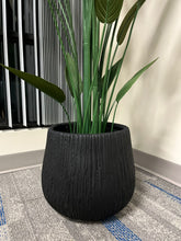 Charger l'image dans la galerie, Close-up of a black textured planter with a ribbed design, housing a tall artificial plant with green stems and leaves, placed indoors near a window. Is this planter made from weather-resistant material?