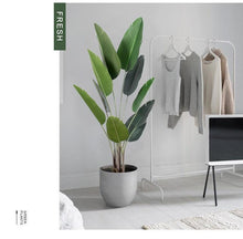 Load image into Gallery viewer, A stylish interior featuring an artificial bird of paradise plant in a sleek gray planter. The plant's large green leaves add a fresh, vibrant touch to the minimalist space. Nearby, a white clothing rack displays neutral-toned sweaters and tops, contributing to the room's clean, modern aesthetic. The muted color palette and simple furnishings create a serene and contemporary environment