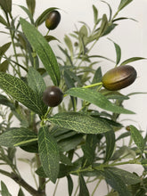 Load image into Gallery viewer, Elegant living room setup featuring a tall artificial olive tree in a white pot positioned next to a modern beige sofa with gold trim. The room has a warm, brown-toned wall adorned with a colorful landscape painting, creating a cozy and sophisticated atmosphere