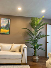 Charger l'image dans la galerie, A tall, artificial palm tree in a woven basket planter placed beside a modern cream-colored couch in a cozy living room. The neutral-toned walls and warm lighting add to the serene and stylish ambiance of the room, complemented by a vibrant wall painting above the sofa.
