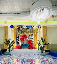 Load image into Gallery viewer, A luxurious event stage decorated with a vibrant setup, featuring a teal loveseat adorned with red floral arrangements and framed by intricate white lattice panels. The backdrop includes cascading gold curtains and an ornate canopy with blue and gold accents hanging from the ceiling. Two large artificial bird of paradise plants in decorative planters flank the stage, adding a touch of greenery to the scene. The floor is covered with a glossy, patterned tile that reflects the colorful ambiance of the space,