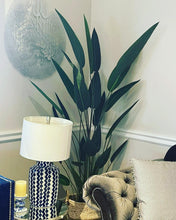Load image into Gallery viewer, A stylish living room corner featuring a tall artificial bird of paradise plant with long, slender leaves placed beside a tufted beige armchair. A decorative blue and white patterned lamp sits on a glass-topped side table, adding a modern touch to the space. The room is accented with wall art and subtle, neutral decor, creating an inviting atmosphere.