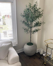 Charger l'image dans la galerie, A cozy living room corner featuring a white sectional sofa with a dark decorative pillow, positioned near a large window with white blinds. To the right of the sofa, a realistic artificial olive tree is placed in a round gray planter on dark hardwood flooring. The corner is accented by a gold-colored bar cart, adding a touch of elegance to the modern, minimalist space. Natural light filters through the window, enhancing the warm and inviting ambiance of the room.