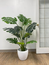 Load image into Gallery viewer, Large artificial monstera plant with vibrant green, split leaves, displayed in a sleek white pot on a wooden floor, enhancing the indoor decor with a tropical touch