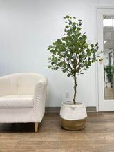 Load image into Gallery viewer, Artificial plant with broad, green leaves displayed in a modern, round white pot, placed next to a beige armchair, adding a natural touch to the indoor setting.
