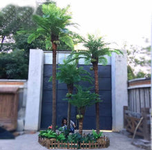 Charger l'image dans la galerie, The image shows a cluster of artificial palm trees with coconuts in a small, fenced garden bed. The setup is outdoors, against a large dark gate, creating a tropical vibe.