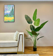 Load image into Gallery viewer, Artificial bird of paradise plant with large green leaves in a black pot placed next to a beige sofa with gold accents in a contemporary living room setting