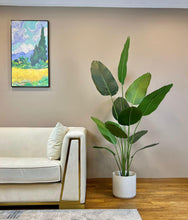 Load image into Gallery viewer, Modern living room with a tall artificial bird of paradise plant in a white pot next to a cream sofa with gold accents and a colorful landscape painting on the wall